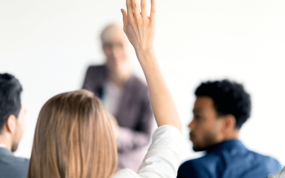 Woman is in a workshop setting, holding her hand up to ask a question as the instructor looks at her.